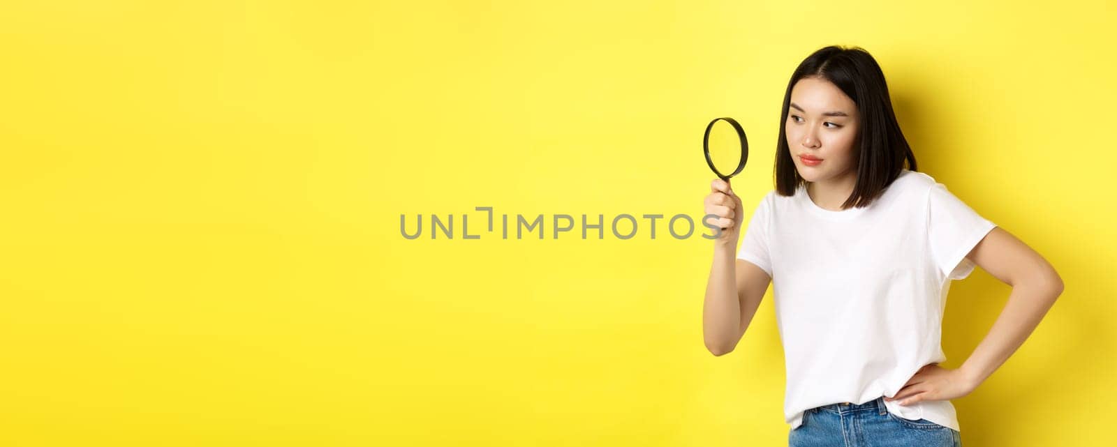 Asian woman detective looking through magnifying glass with intrigued look, found clues, standing over yellow background by Benzoix