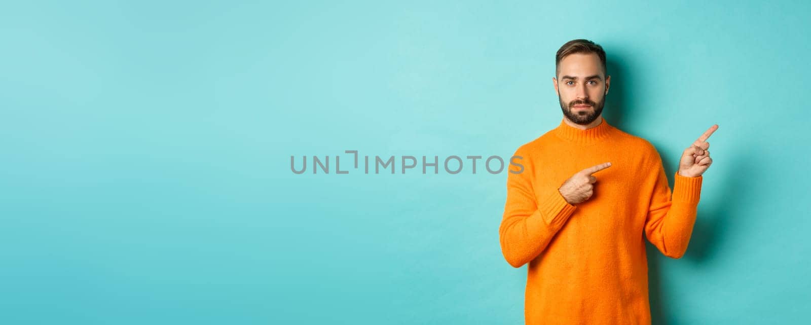 Handsome and serious bearded man in orange sweater pointing right, showing advertisement or logo, standing over turquoise background.