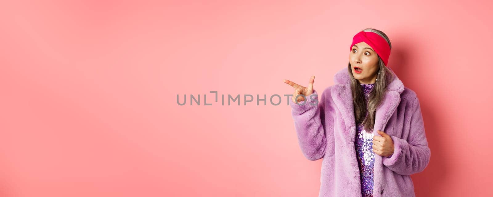 Fashion and shopping concept. Trendy elderly asian female in fake fur and glittering dress pointing, looking left with amazement, checking out offer, pink background.