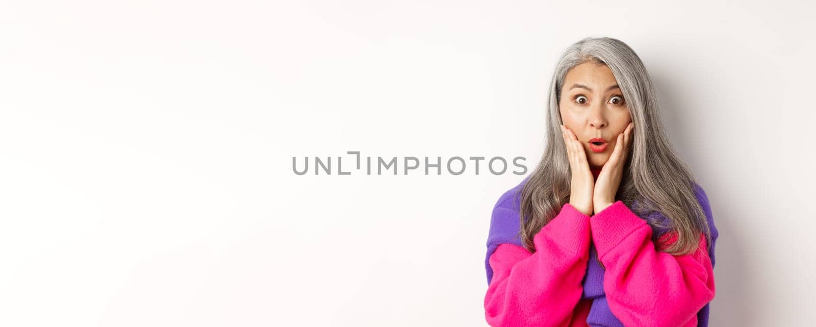 Close up of fashionable asian senior woman with grey hair looking surprised, saying wow and staring at camera amazed, standing over white background by Benzoix