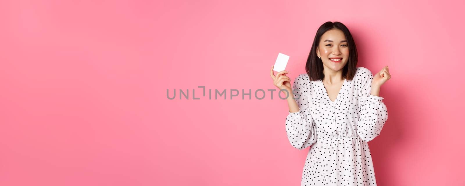 Shopping concept. Pretty asian woman holding bank credit card and smiling, standing over pink background by Benzoix
