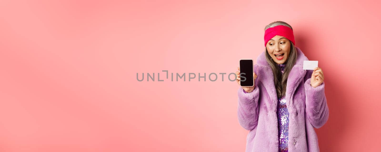 Special promotion. Excited asian senior woman showing blank smartphone screen and plastic credit card, checking out online offer, pink background by Benzoix