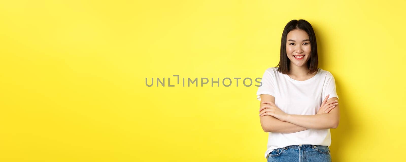 Confident and stylish asian woman cross arms on chest and smiling, standing over yellow background.