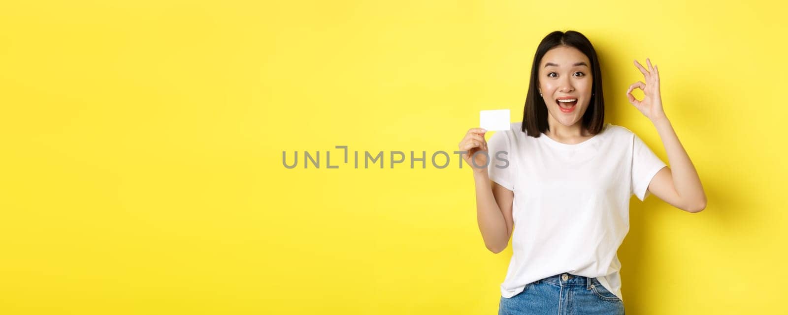 Young asian woman in casual white t-shirt showing plastic credit card and okay gesture, recommend bank, smiling at camera, yellow background.