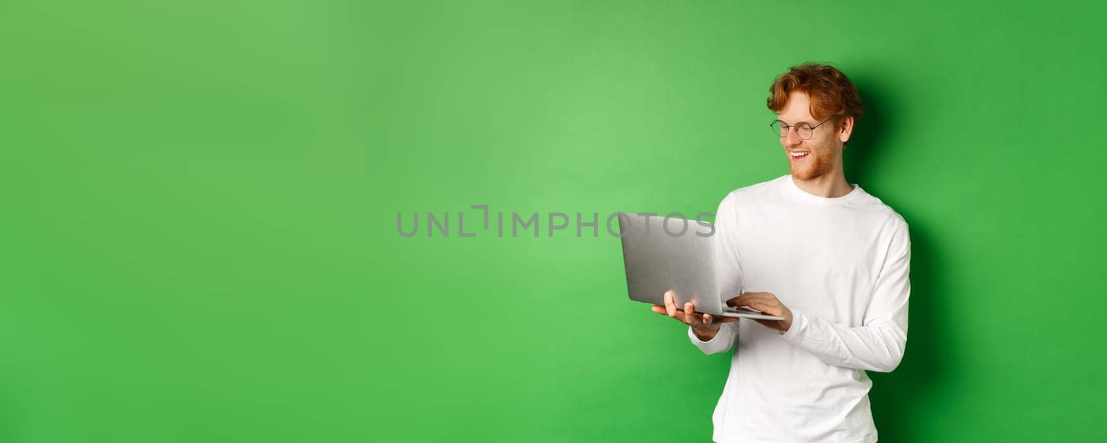 Smiling young man with red hair, wearing glasses, working on laptop and smiling, standing over green background by Benzoix
