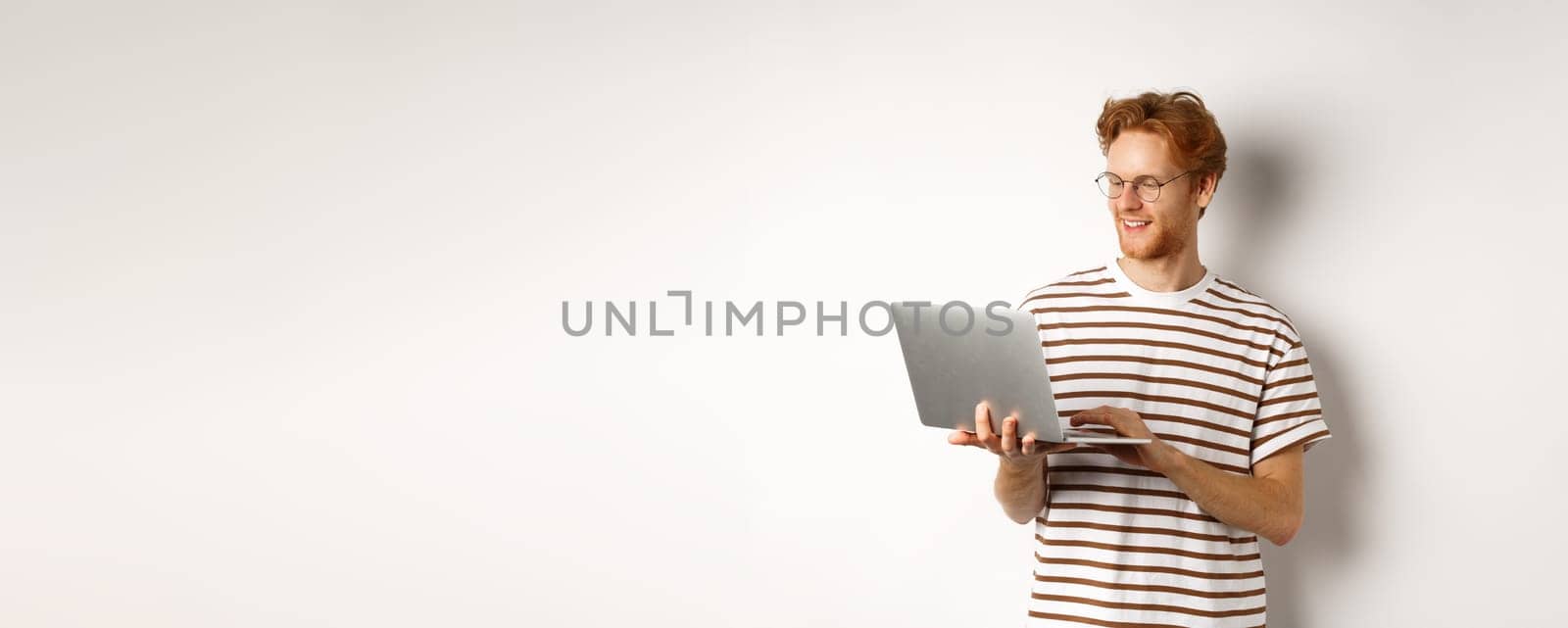 Young redhead freelancer working on laptop, typing on computer keyboard and smiling, standing over white background.