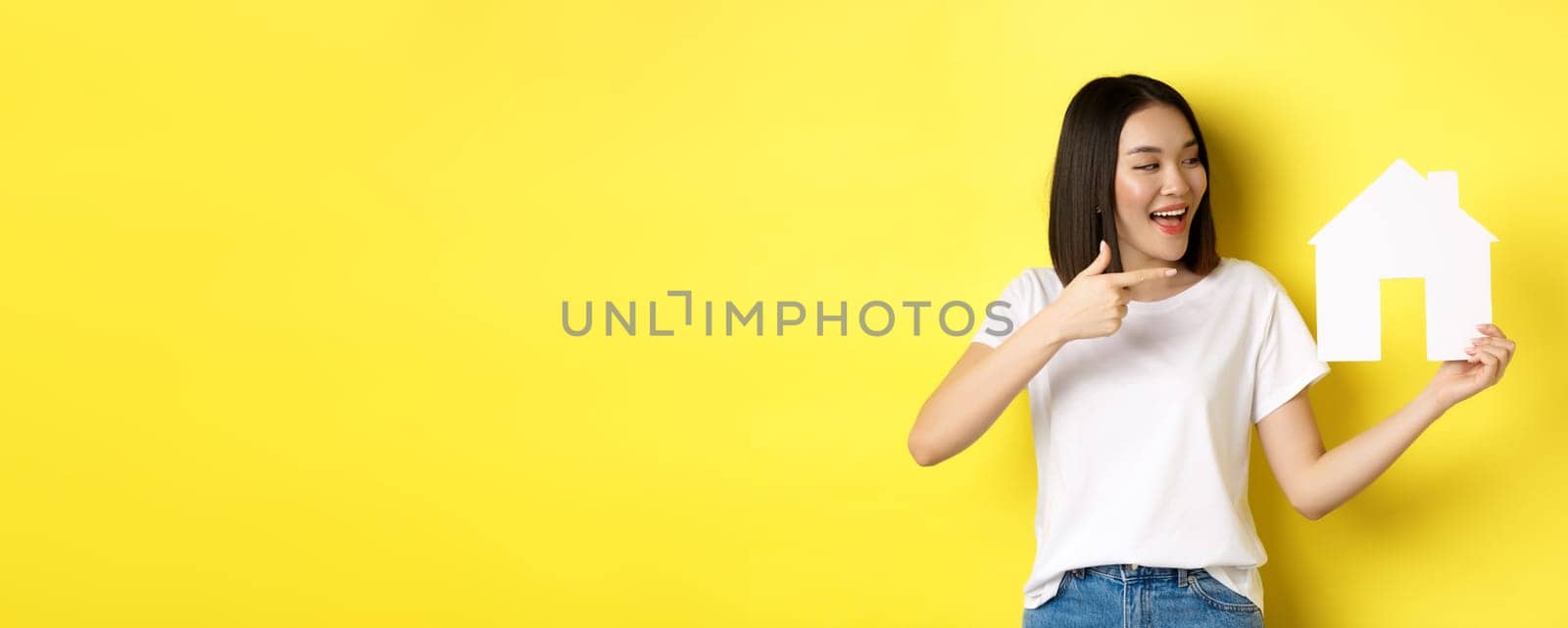 Real estate and insurance concept. Cheerful asian woman smiling, pointing and looking at paper house cutout, recommend agency logo, standing over yellow background.