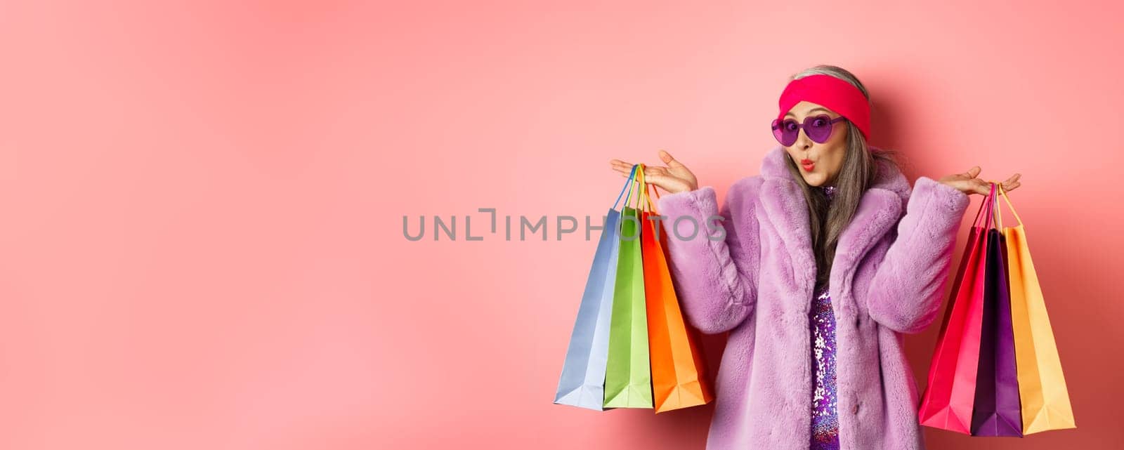 Stylish asian senior woman going shopping, wearing trendy clothes and sunglasses, holding store bags with clothes and purchases, pink background.