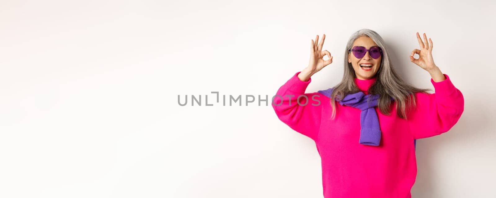 Stylish modern asian grandmother in sunglasses and pink sweater, smiling happy and showing OK signs, approve something cool, white background.