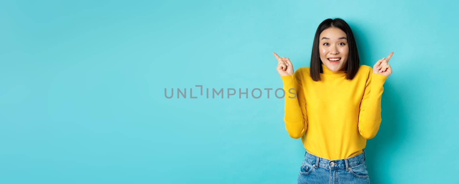 Beauty and fashion concept. Cheerful asian girl showing two promo offers, pointing fingers sideways at left and right advertisements and smiling, standing over blue background.