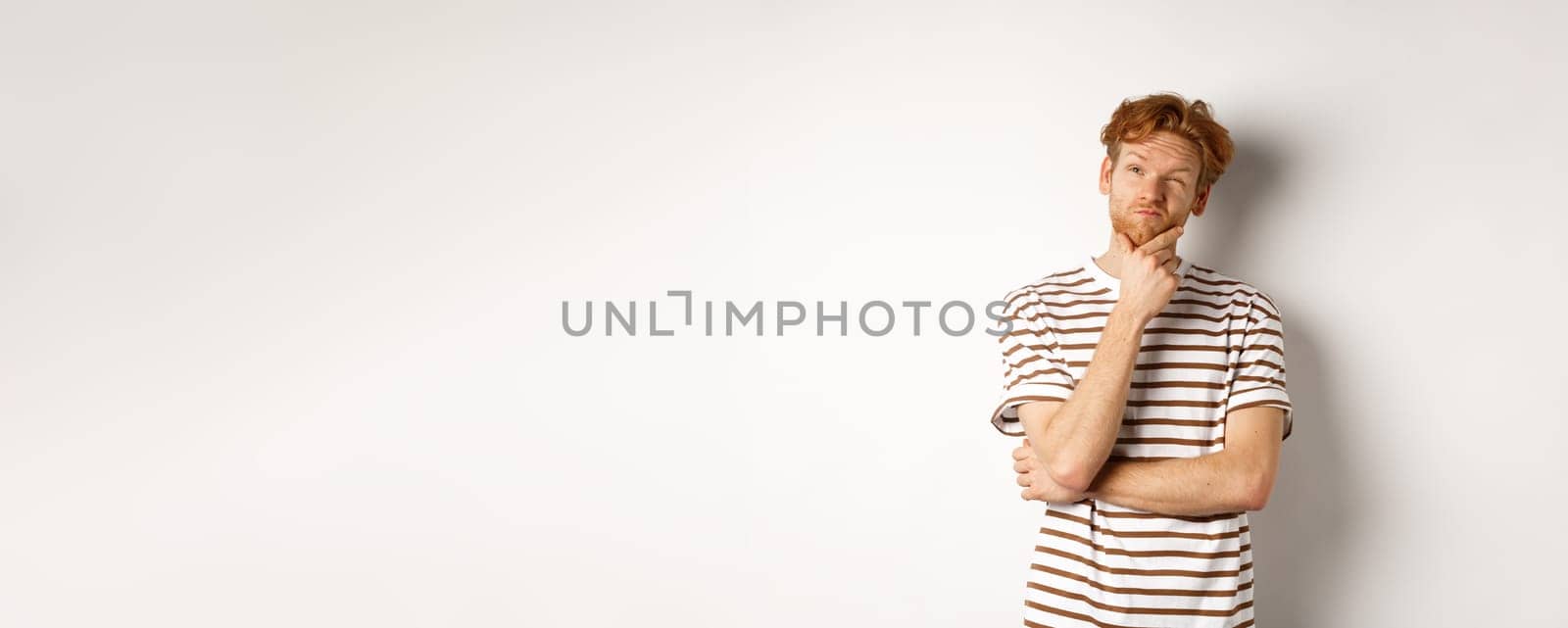 Portrait of thoughtful man with red hair thinking, looking at upper left corner and making choice, standing over white background by Benzoix