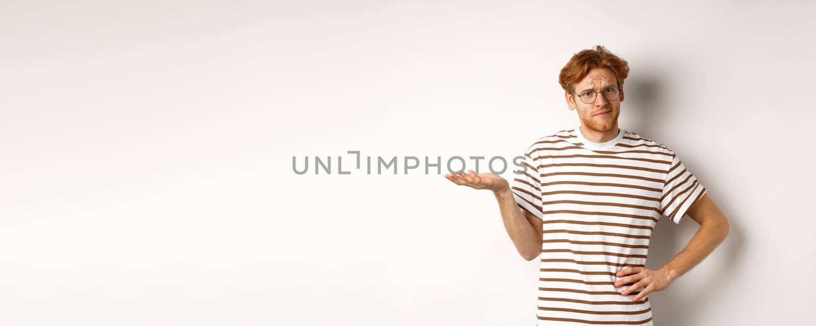 So what. Confused and annoyed redhead man waiting for reply, standing questioned with raised hand, holding something on palm, white background.