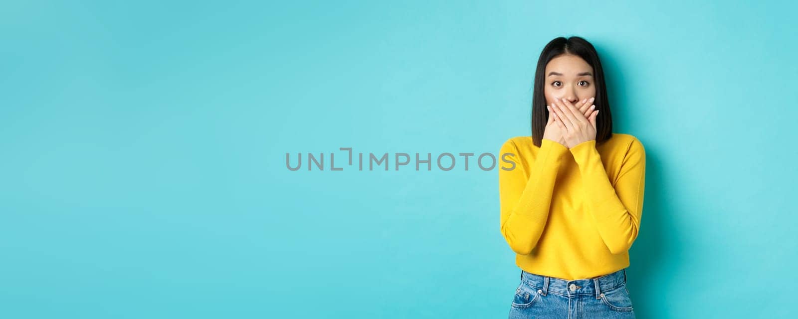 Image of shocked asian girl cover mouth and staring at camera, wearing yellow sweater against blue background.