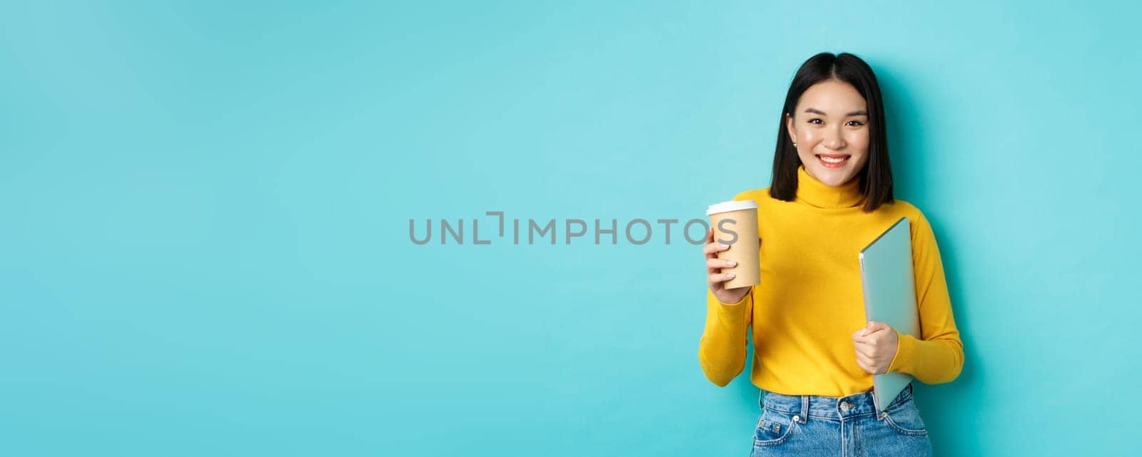 Stylish asian female student with cup of coffee standing over blue background, holding laptop in hand, smiling at camera, standing over blue background.