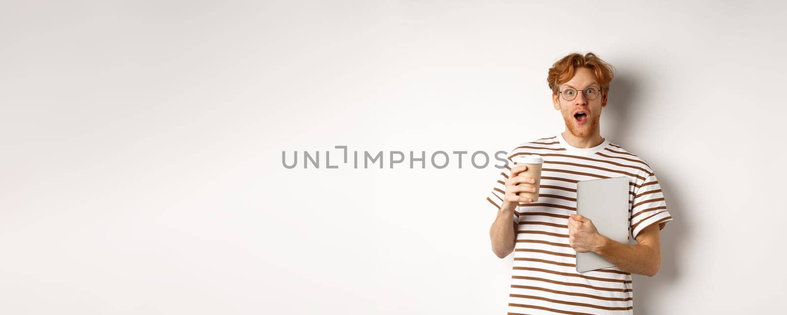 Amazed redhead man talking with coworkers on coffee break, holding cup and laptop, staring surprised at camera, white background.