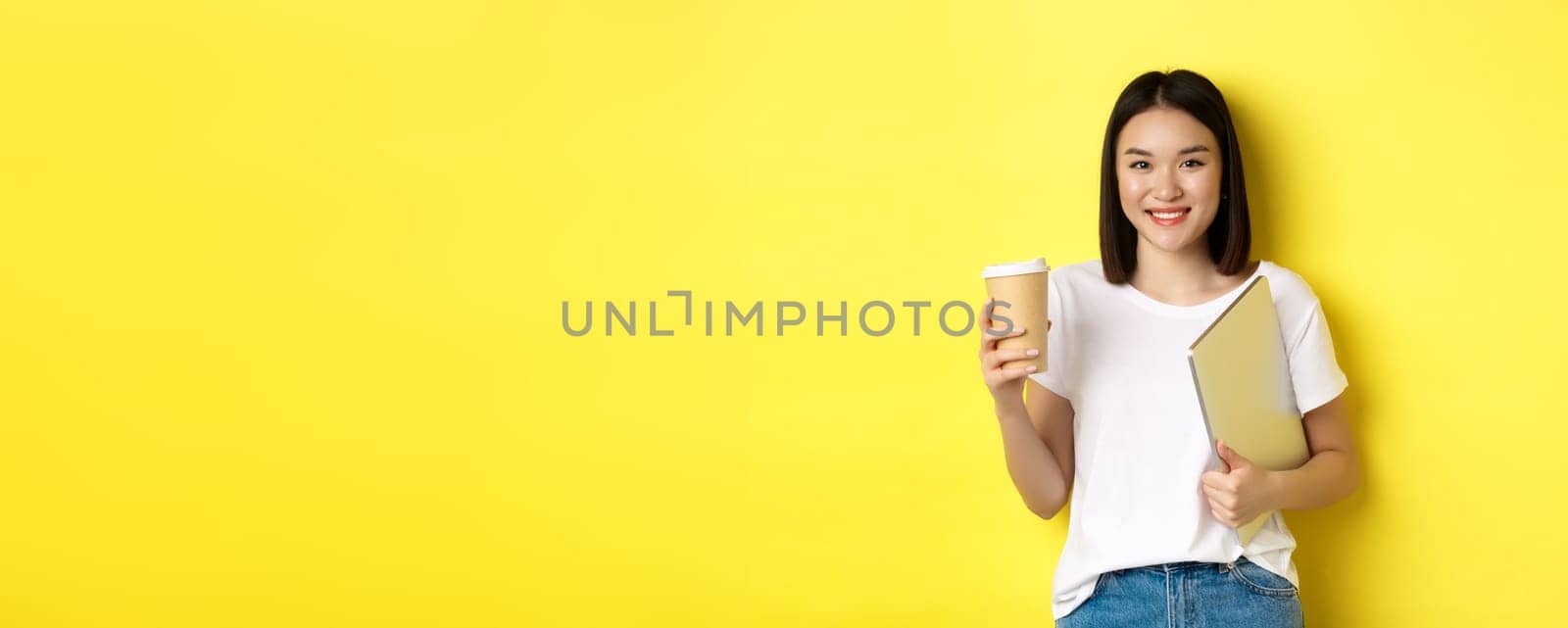 Asian girl student drinking coffee and holding laptop, smiling at camera, standing over yellow background by Benzoix