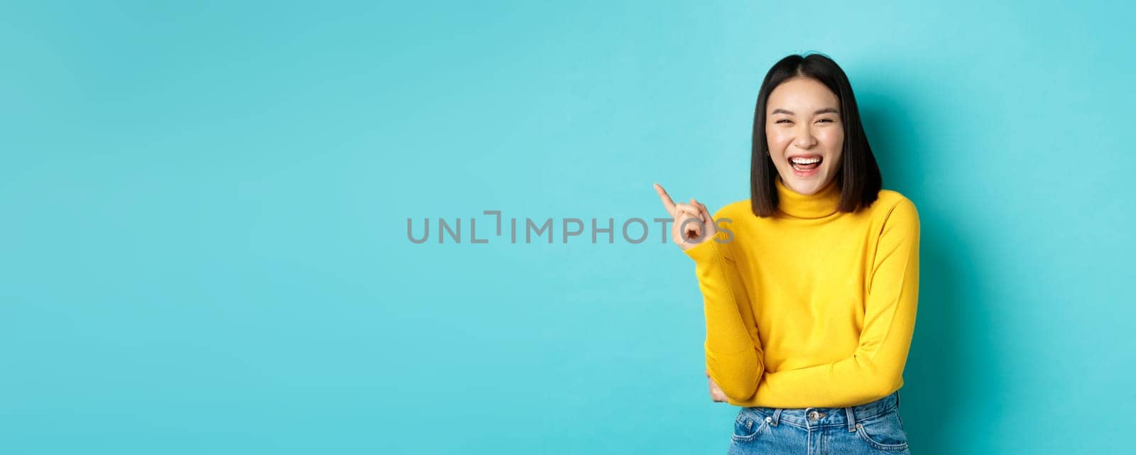 Image of carefree pretty korean girl laughing and looking happy, pointing finger at upper left corner promo, standing against blue background.