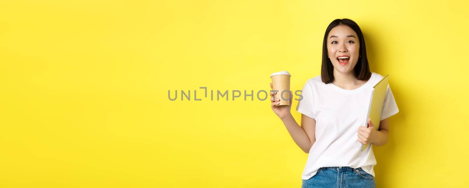 Happy asian woman drinking coffee and holding laptop, saying hi to you, standing over yellow background.