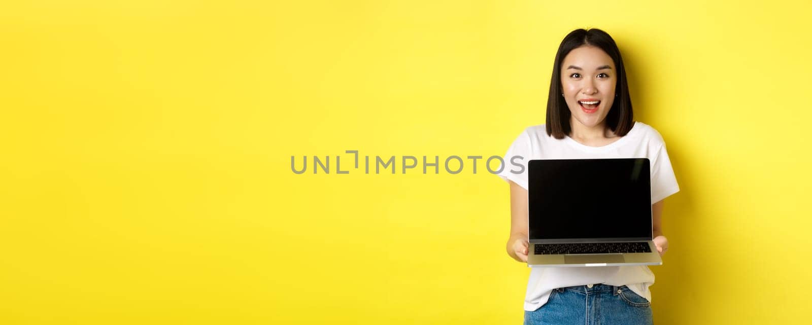 Young asian woman demonstrate online offer, showing blank laptop screen and smiling, standing over yellow background by Benzoix