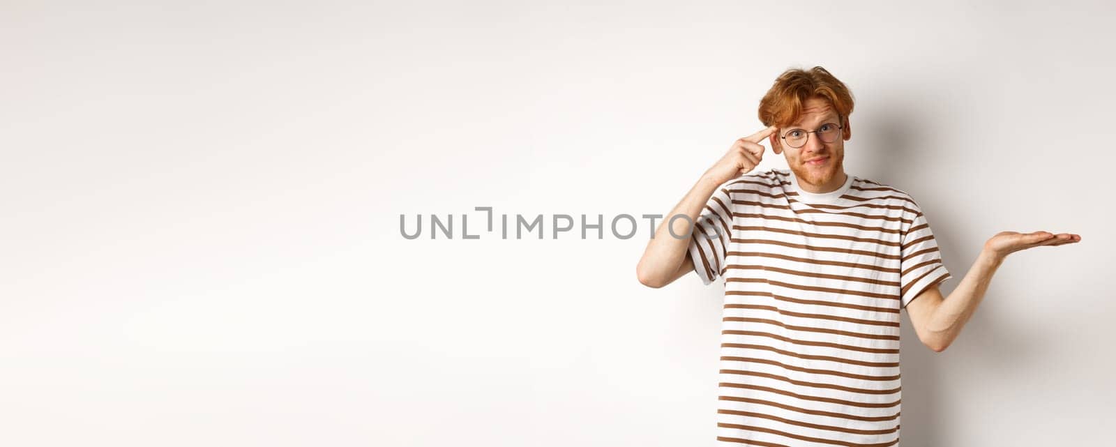 Are you stupid. Confused redhead guy in glasses shrugging, pointing at head and staring skeptical at camera, white background.