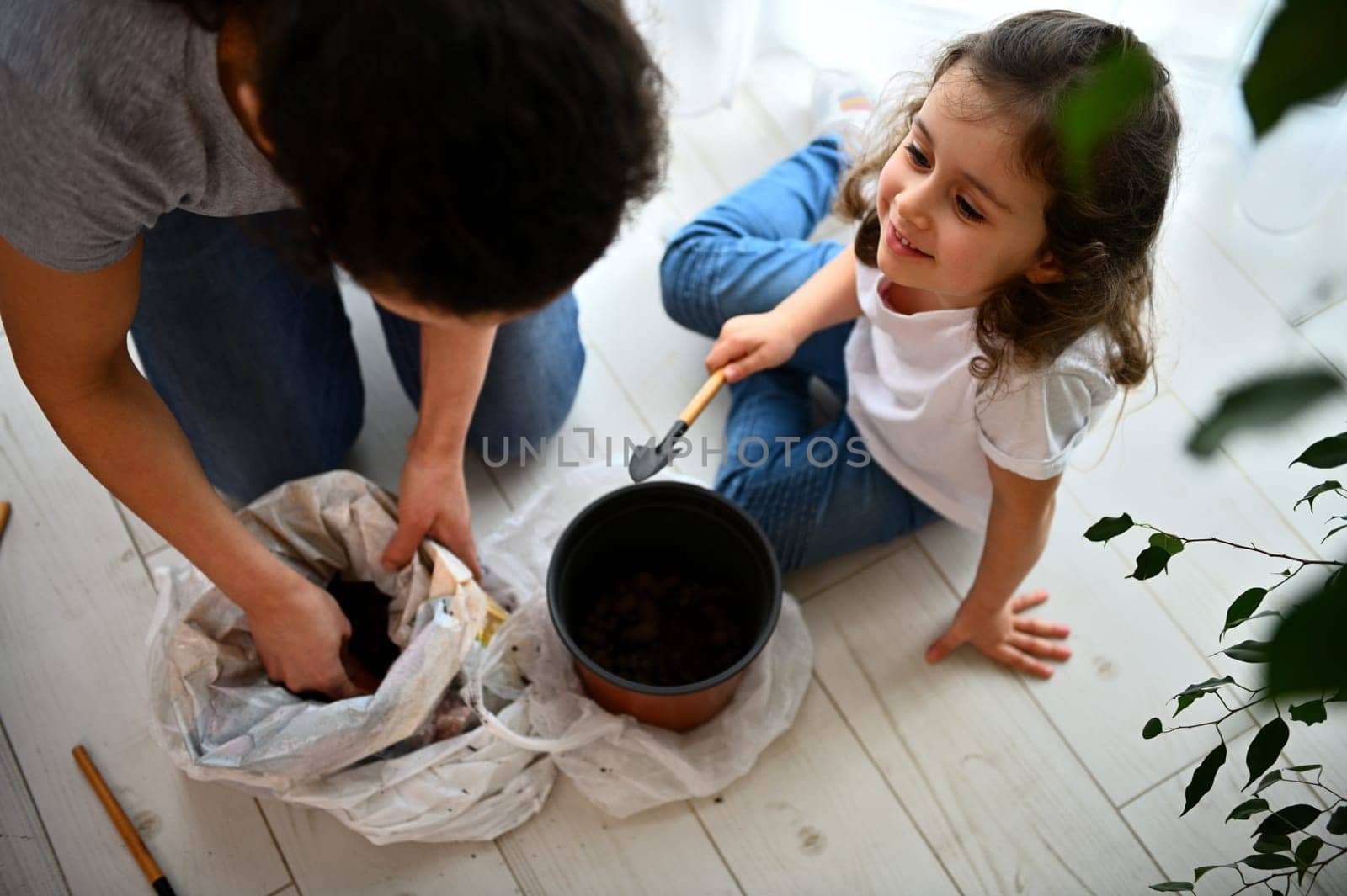 Little girl sitting on floor with garden shovel in her hand, smiling to her mom working with soil white repotting plants by artgf