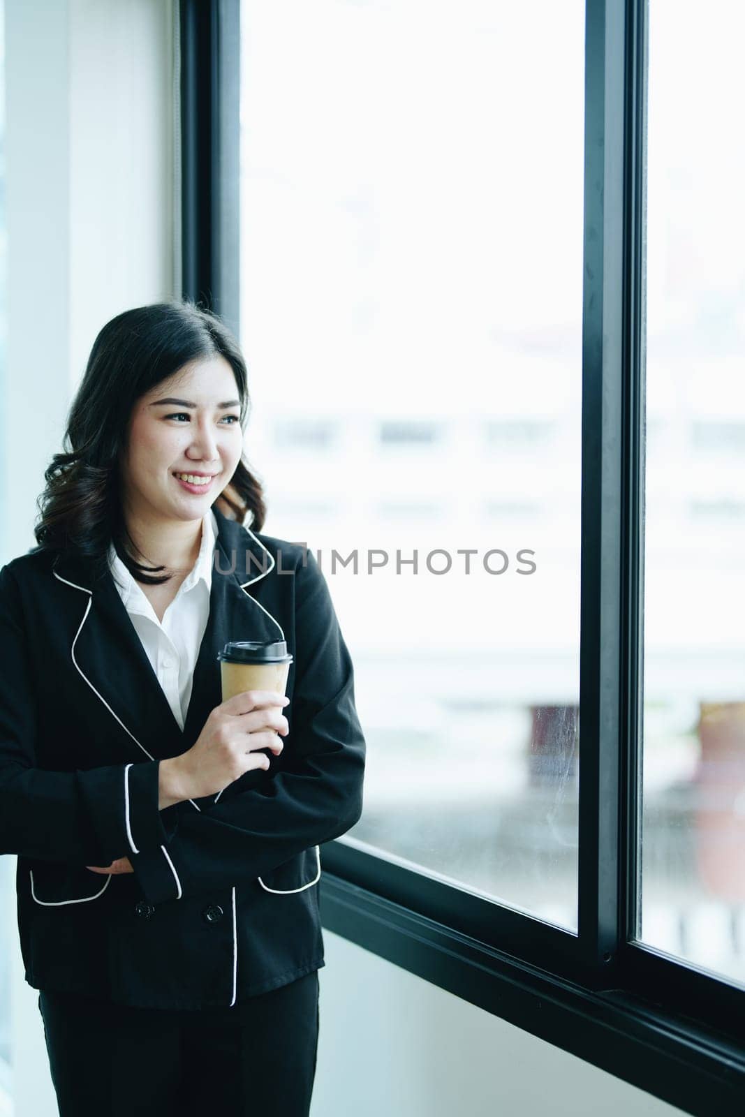 A portrait of a young Asian businesswoman smiling happily and drinking coffee by the window in the conference room.