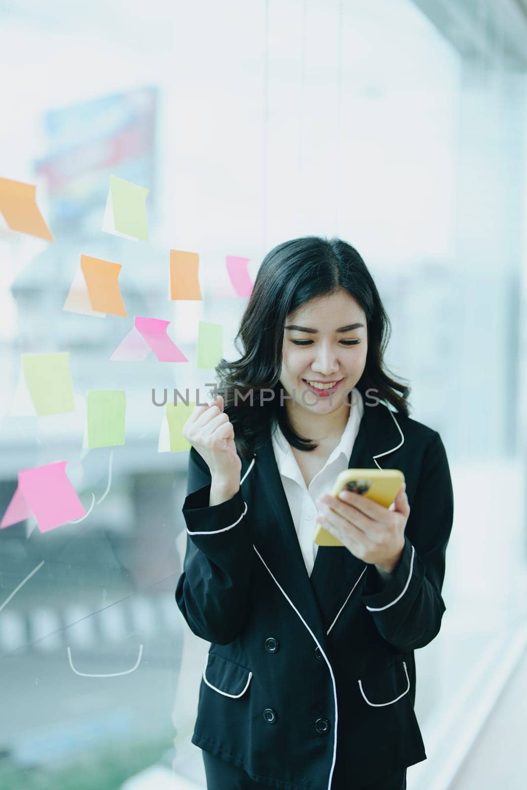 A portrait of an Asian business owner showing a smiling and happy face using her phone standing by a window in a conference room.