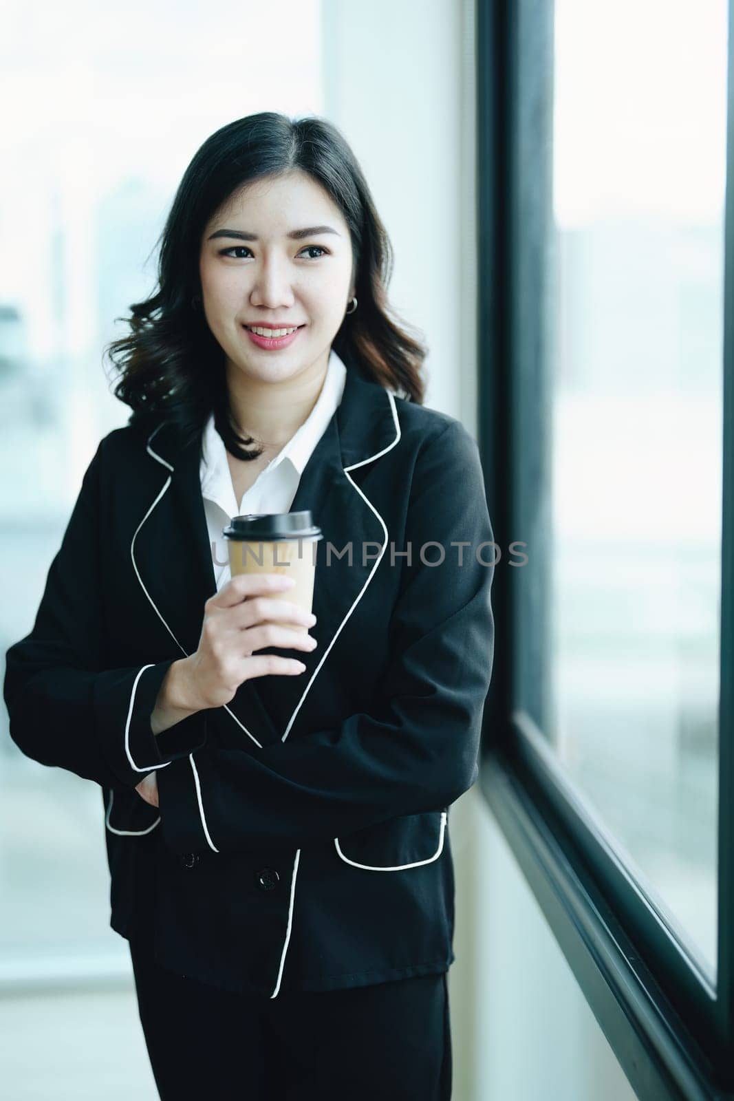 A portrait of a young Asian businesswoman smiling happily and drinking coffee by the window in the conference room.