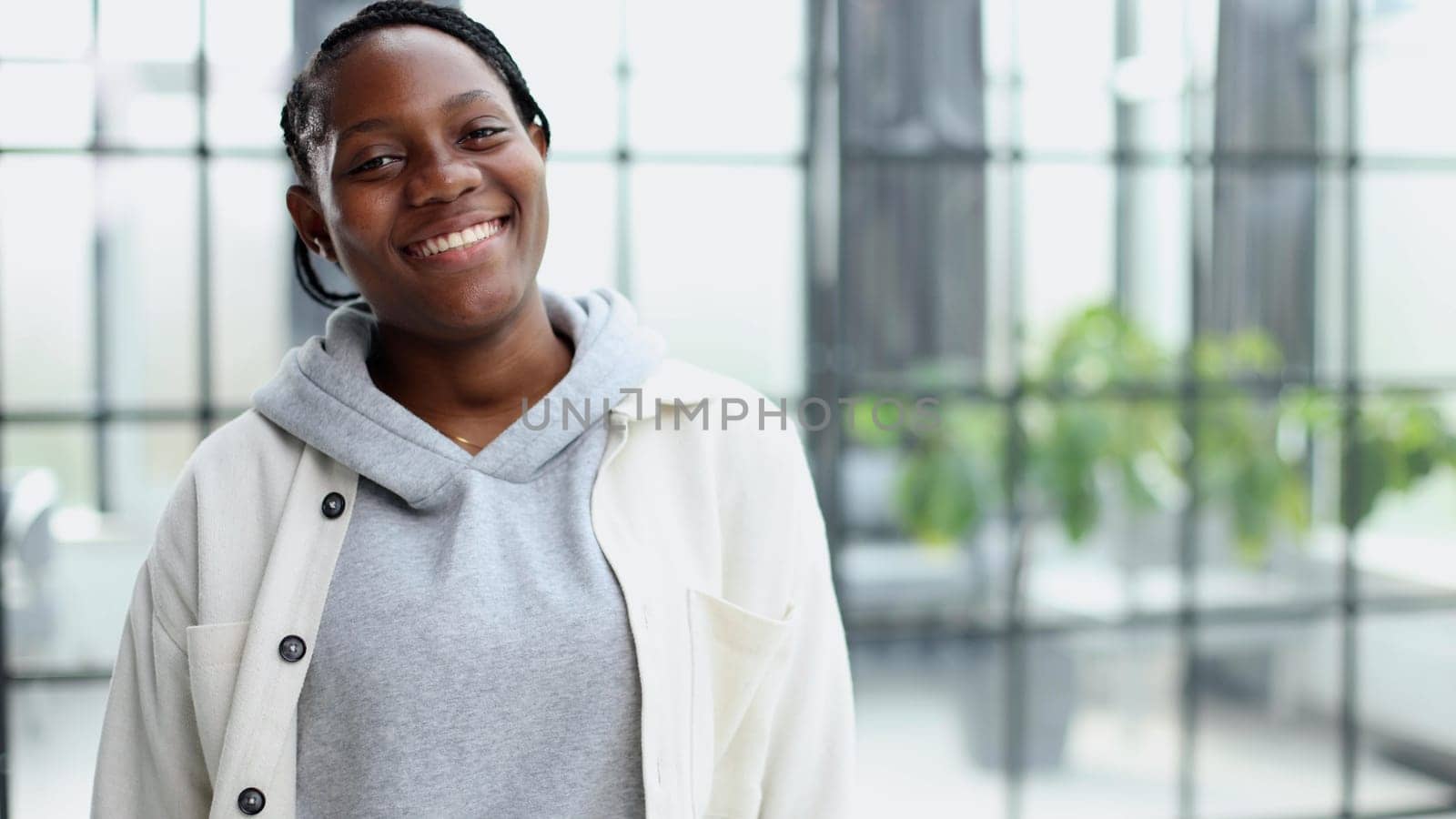Smiling young woman standing and looking at the camera. She stands in a modern office against the background of a window.