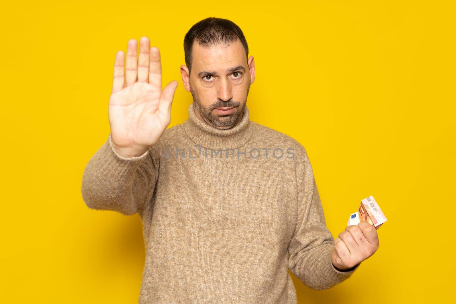 Hispanic man about 40 years old holding a euro bills isolated on yellow background standing with outstretched hand showing the stop sign, preventing you.