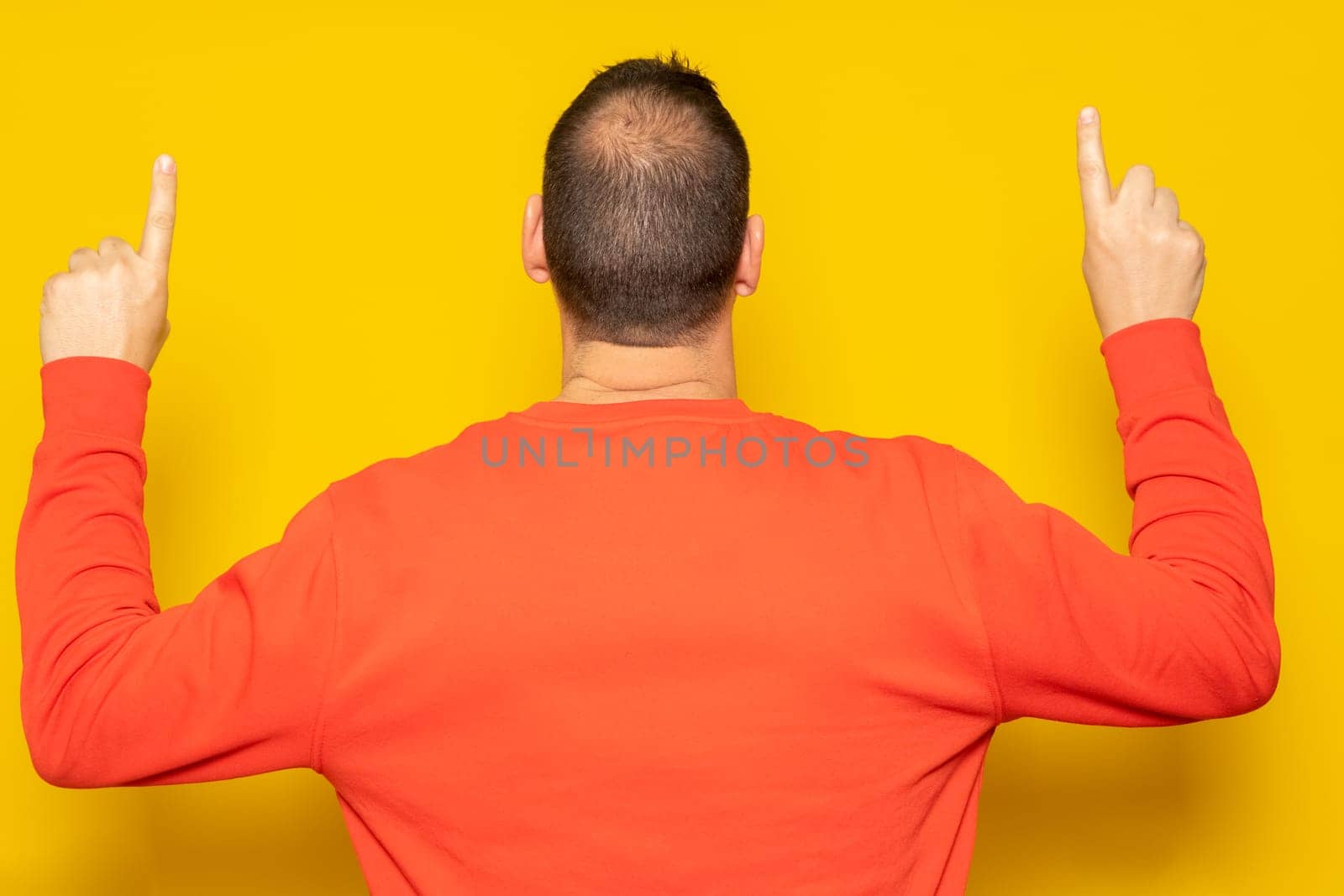 Hispanic man in red sweatshirt pointing up with his index fingers with his back to the camera, isolated on yellow background