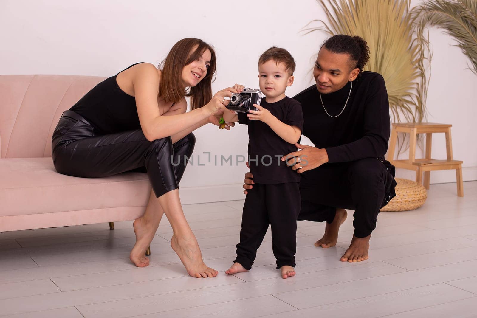 Happy multiracial family with little boy, in black clothes, in the light living room, playing with an old camera