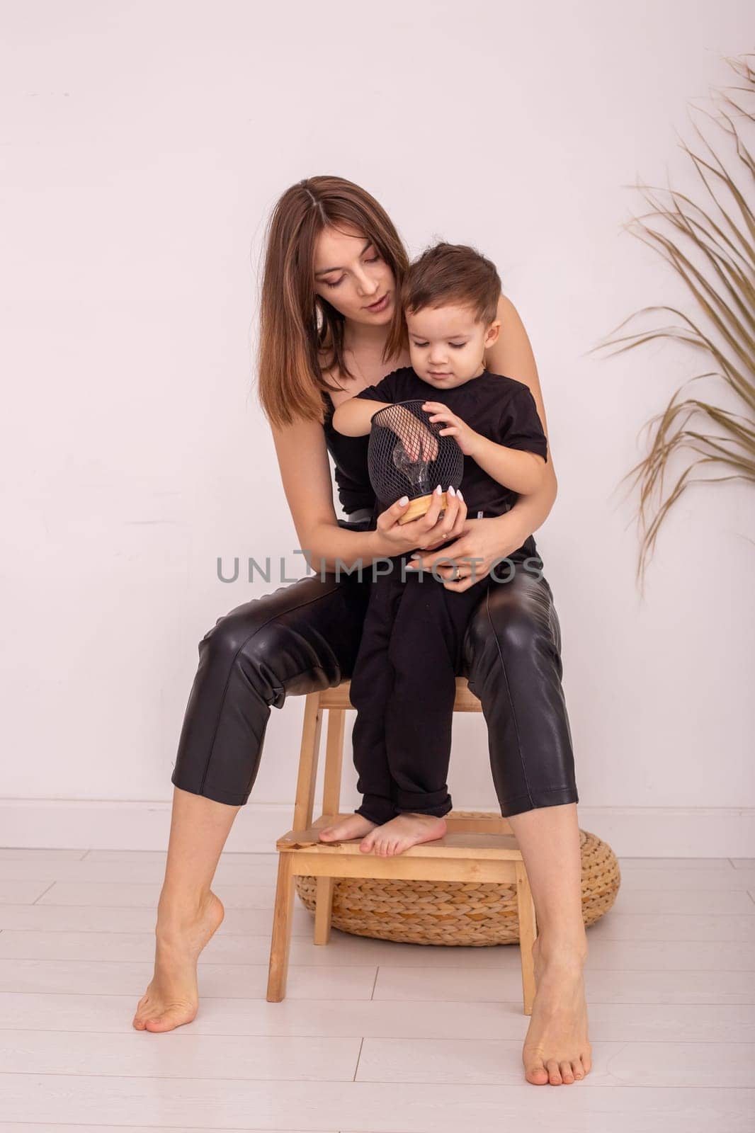 A slender mother with a little boy, in black clothes, is sitting next to a light wall, indoors, hold a lamp. Vertical . Copy space