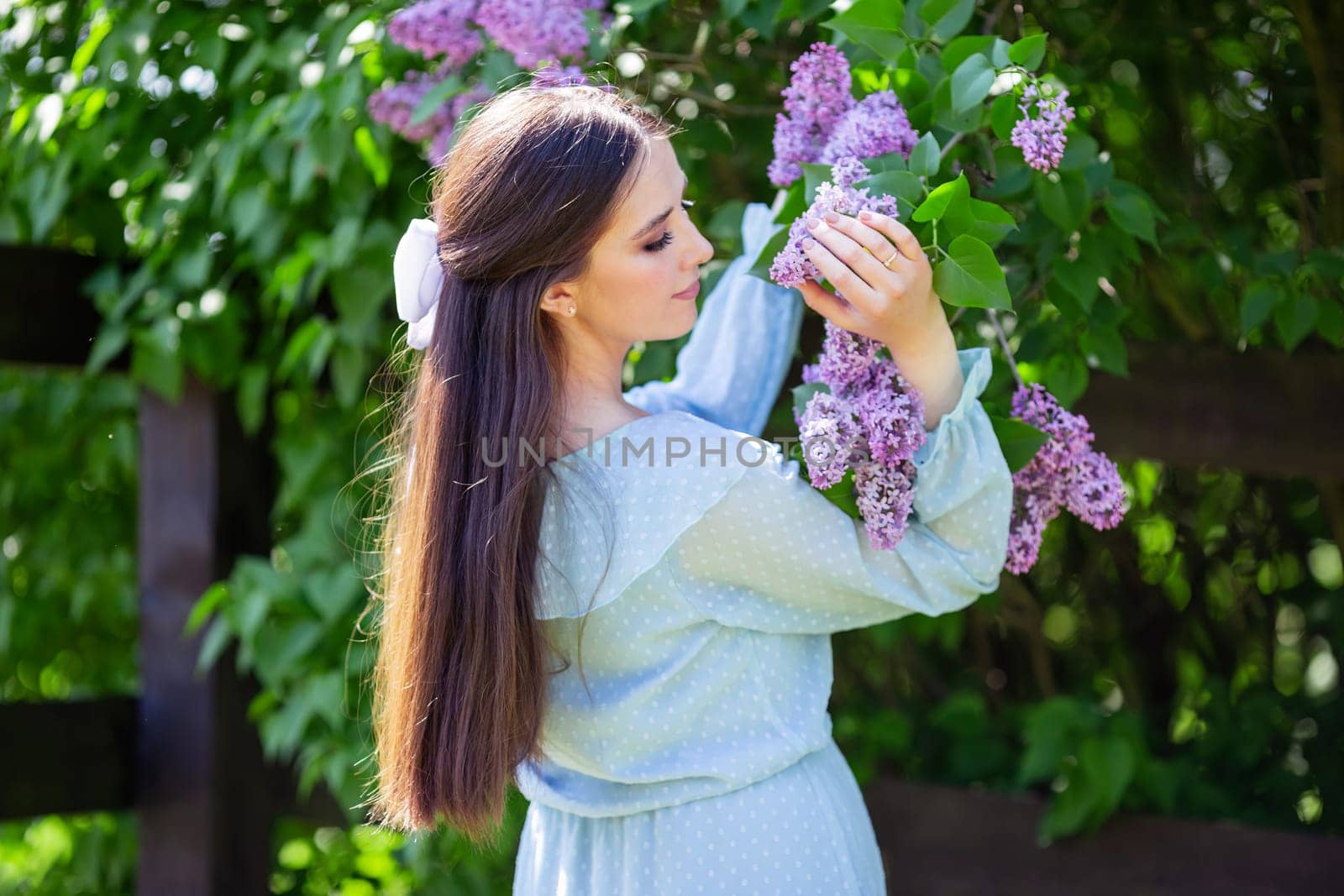romantic girl with long hair with white bow, in light blue dress stands with lilac flowers, in the garden, in sunny day. Close up. copy space