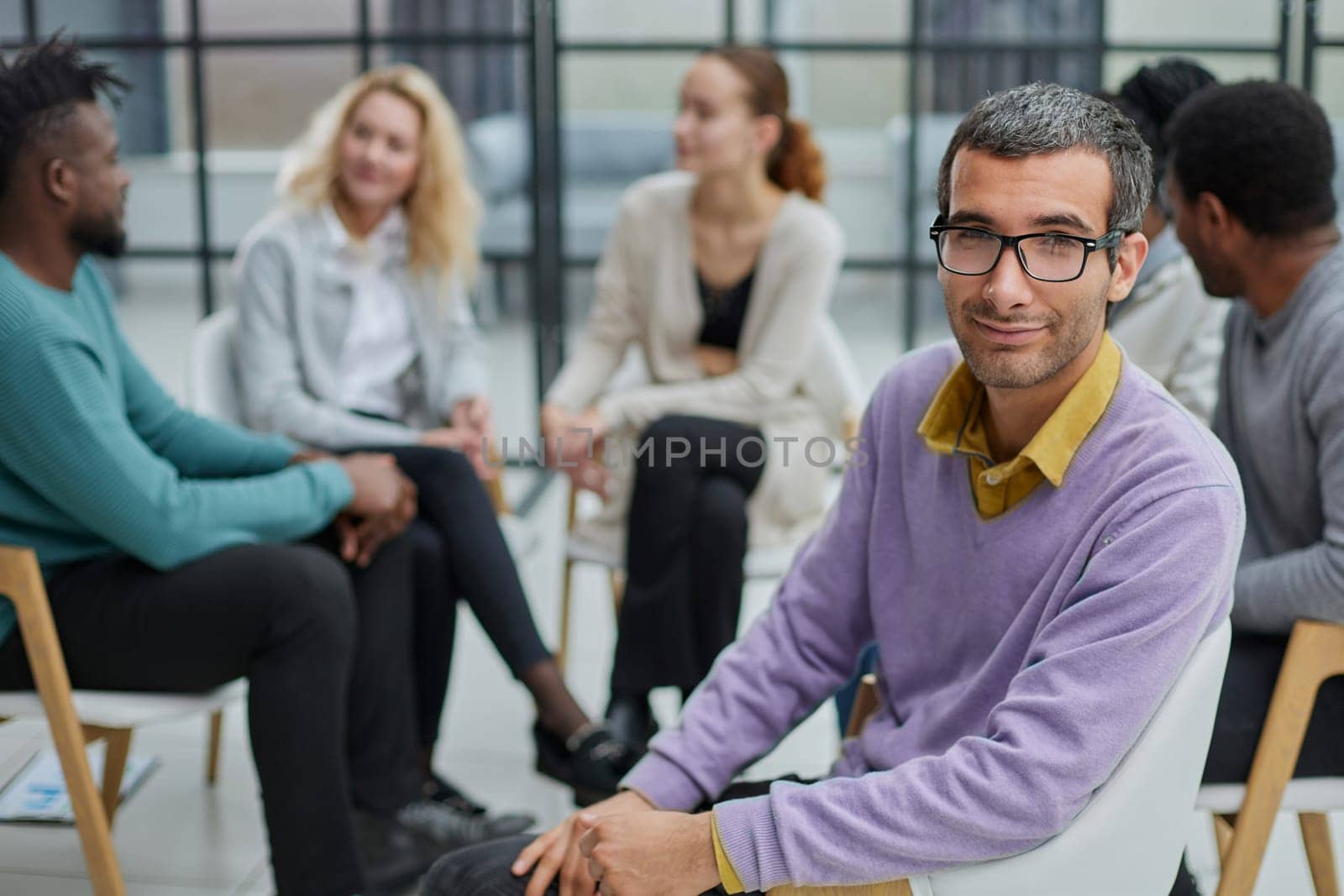 A happy young businessman, in a purple sweater, sits against the background of his colleagues in a modern office
