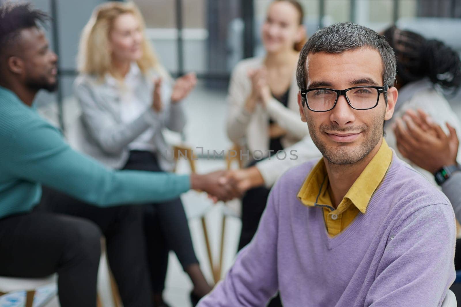A happy young businessman, in a purple sweater, sits against the background of his colleagues in a modern office