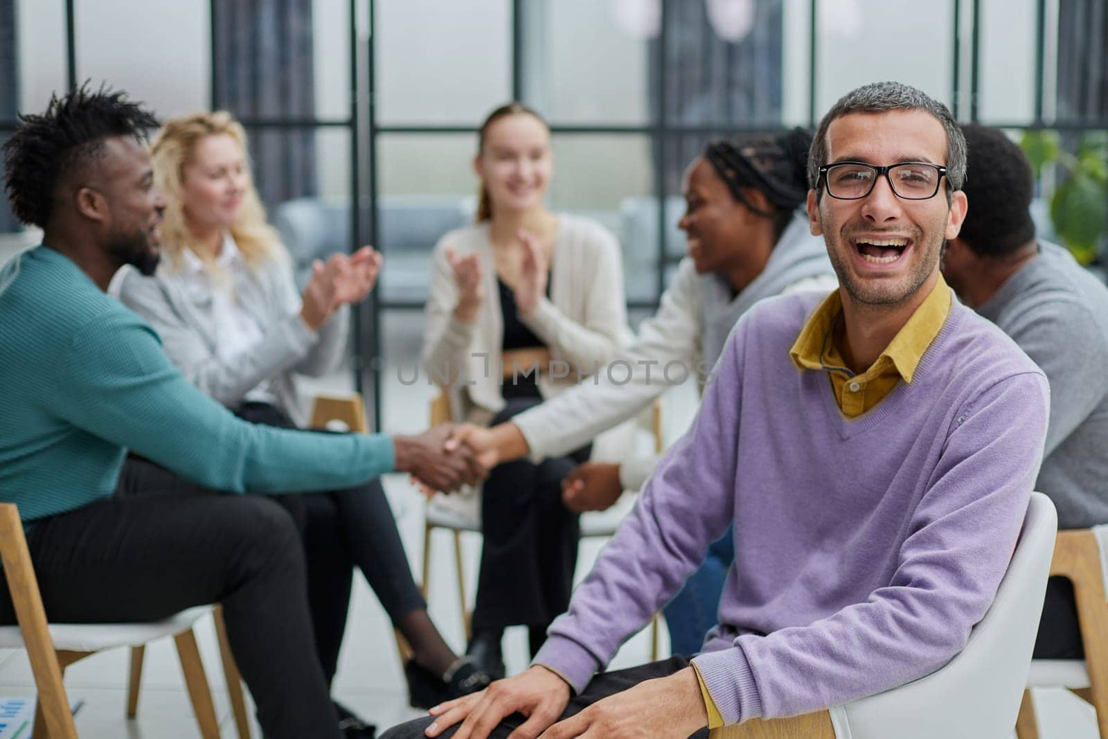 A happy young businessman, in a purple sweater, sits against the background of his colleagues in a modern office