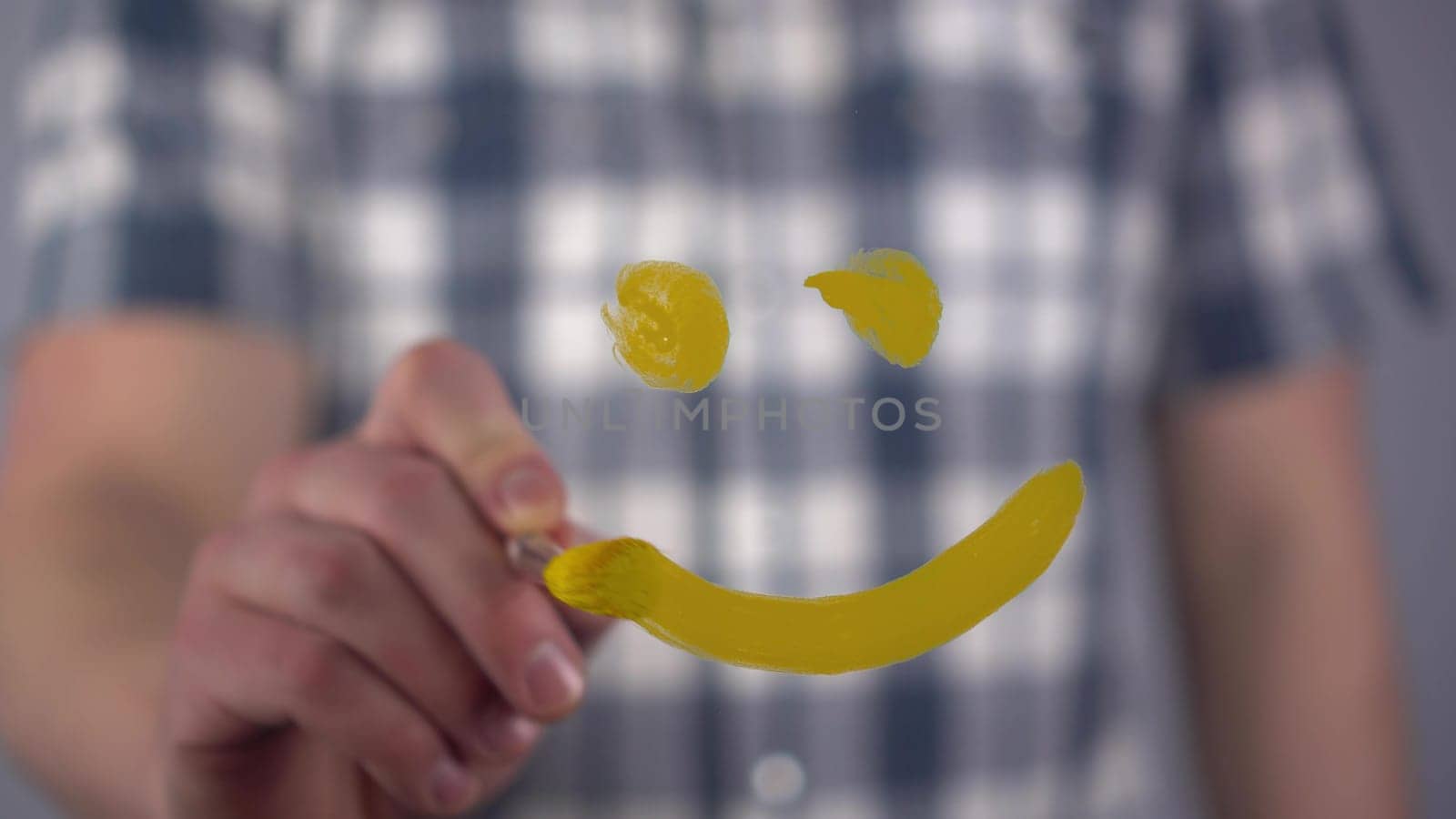 A young man draws a happy smile on the glass. A man in a plaid shirt draws with a brush and yellow paint. Close-up. 4k