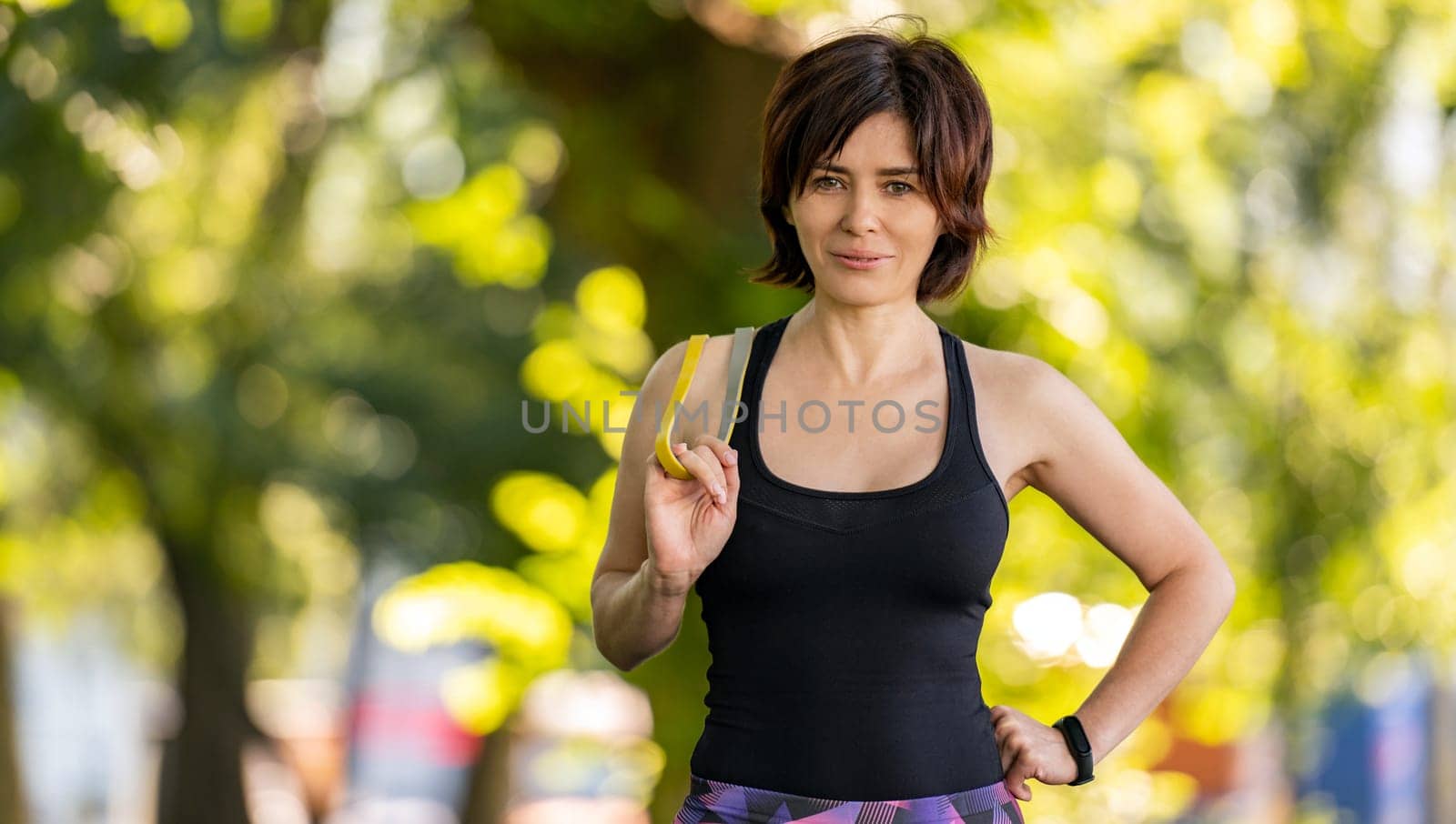 Girl with rubber elastic band looking at camera after workout outdoors in park. Young woman exercising at morning with additional equipment portrait