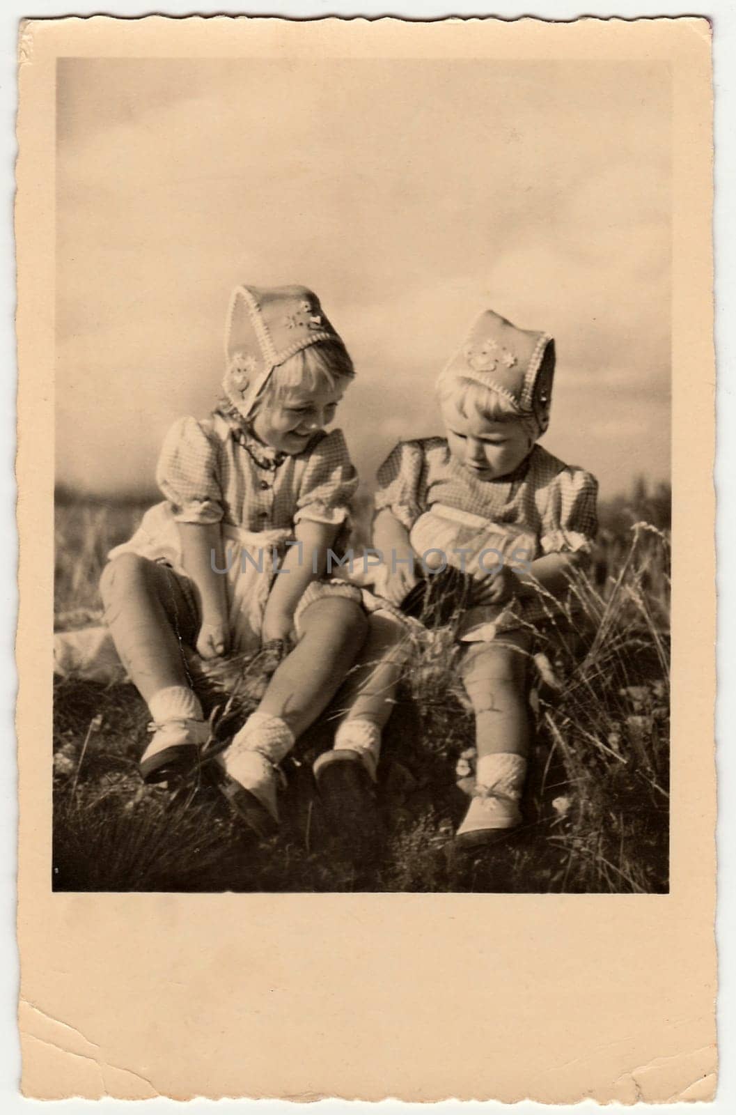 DRESDEN, GERMANY - CIRCA 1954: Vintage photo shows girls sit on grass. They wear traditional cap for girls. Antique black white photo.