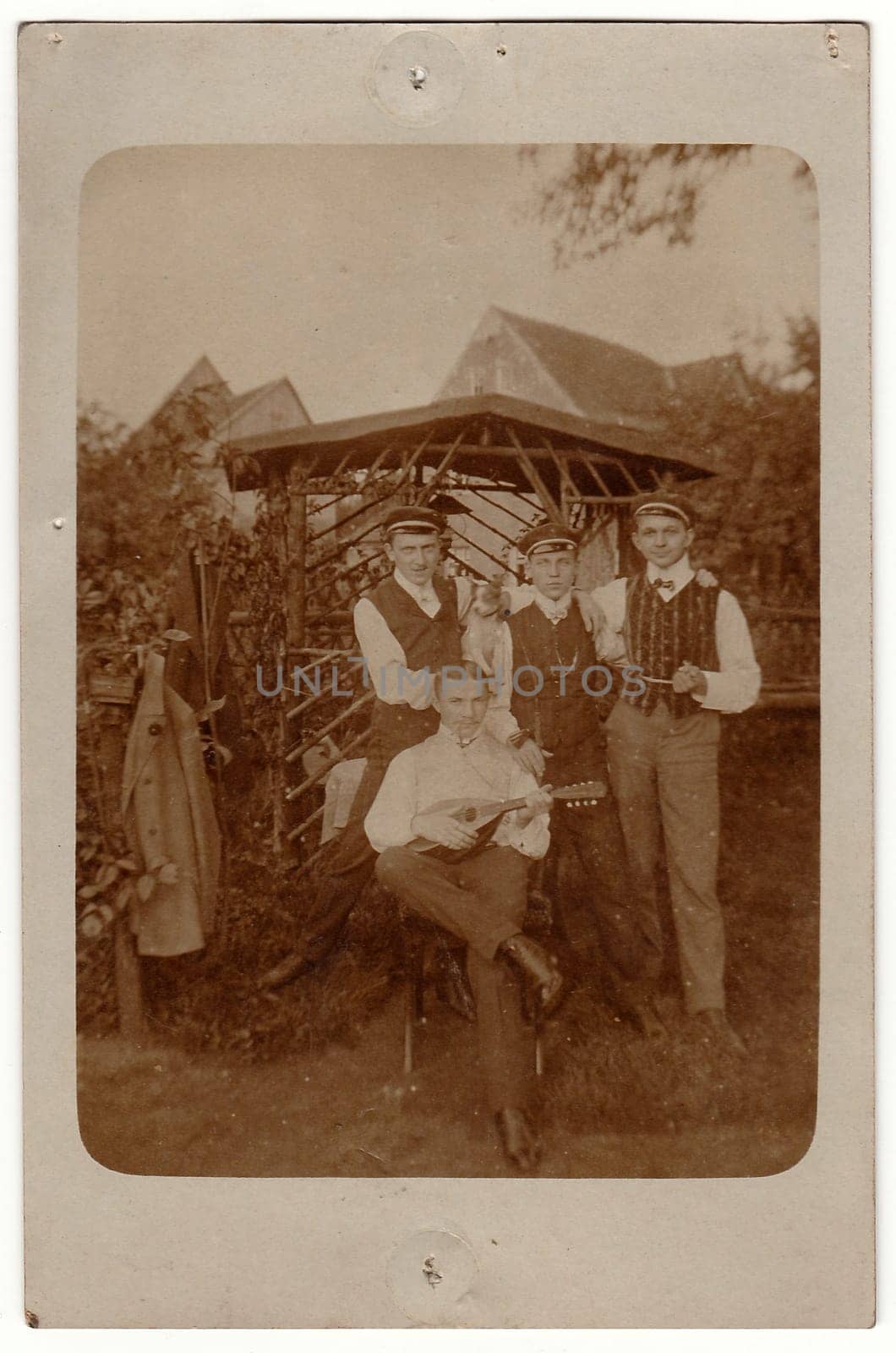 Vintage photo shows men in front of summerhouse. One of them plays the mandolin. Antique black white photo. On top and bottom of image are imprints of drawing pins. by roman_nerud