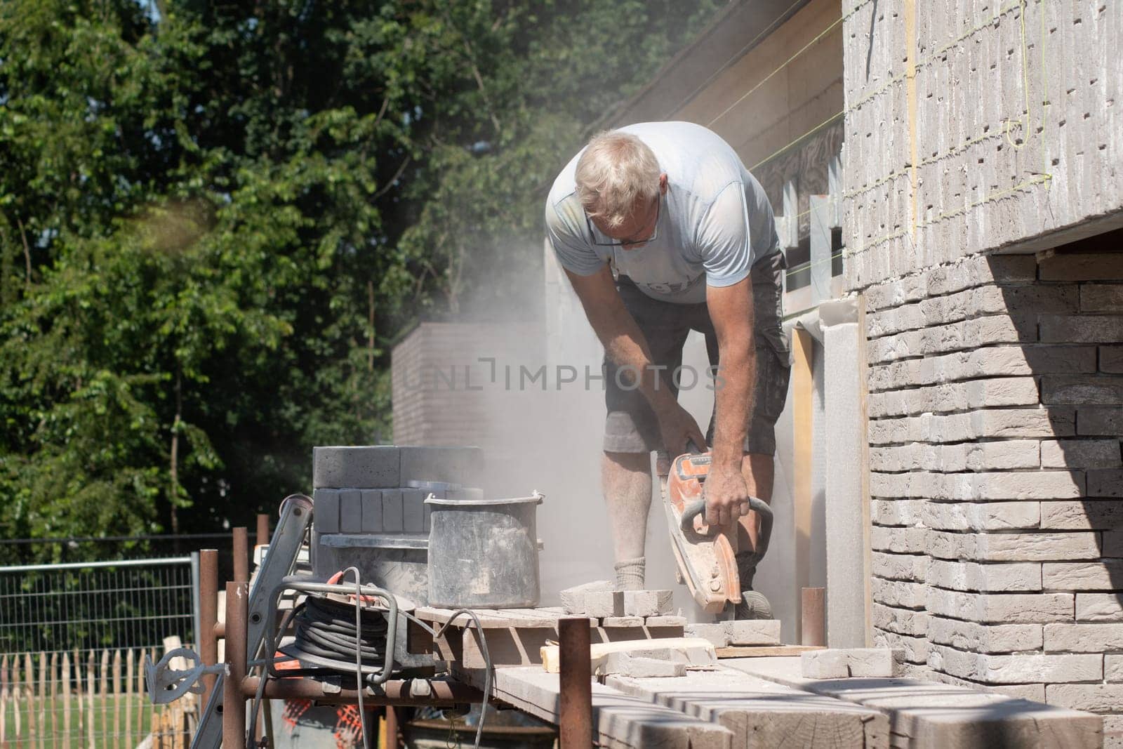 A gray-haired man saw bricks with a concrete saw of the right size for subsequent laying out in the facade of the house, High quality photo