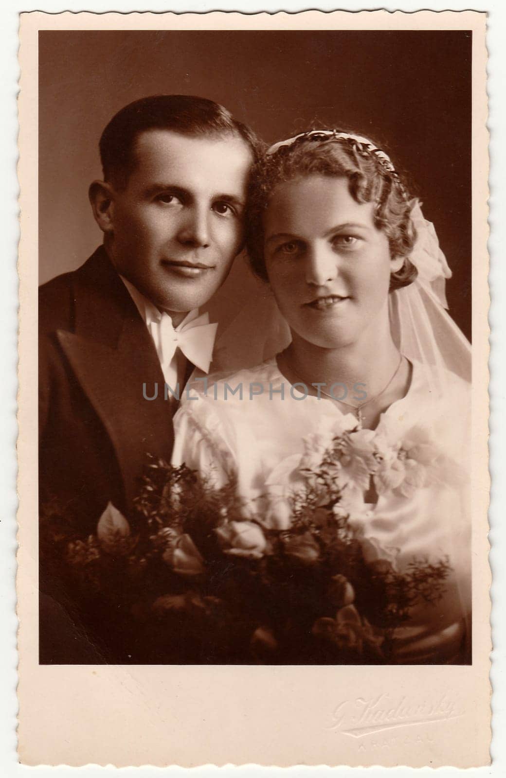 Vintage photo of newlyweds with wedding bouquet. Bride wears wedding veil headdress. Groom wears posh clothing, white bow-tie. Black white antique studio portrait. by roman_nerud