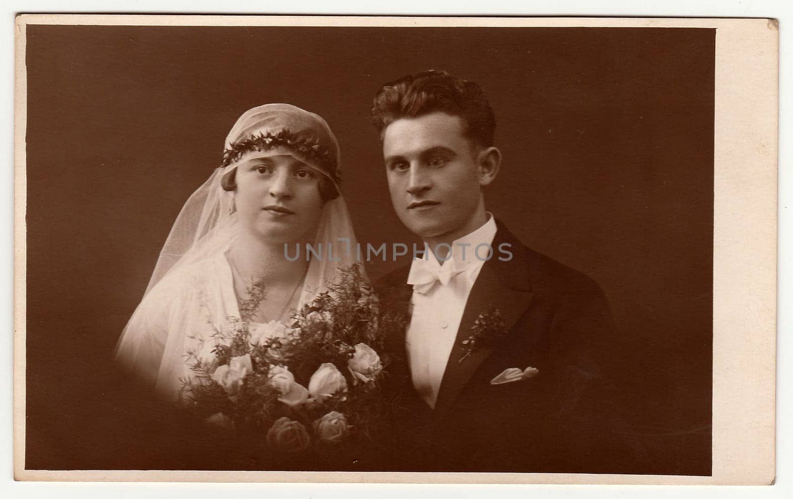 Vintage photo of newlyweds with wedding bouquet. Bride wears wedding veil headdress. Groom wears posh clothing, white bow-tie. Black white antique studio portrait. by roman_nerud