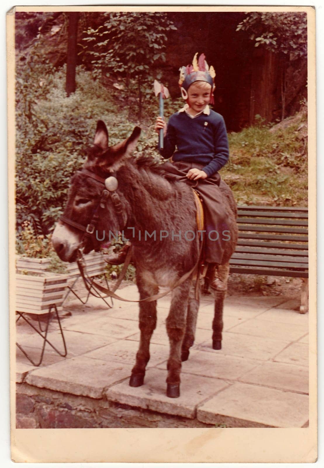 Vintage photo shows boy sits on donkey, he wears Indian headband and holds tomahawk. Colour photography. by roman_nerud