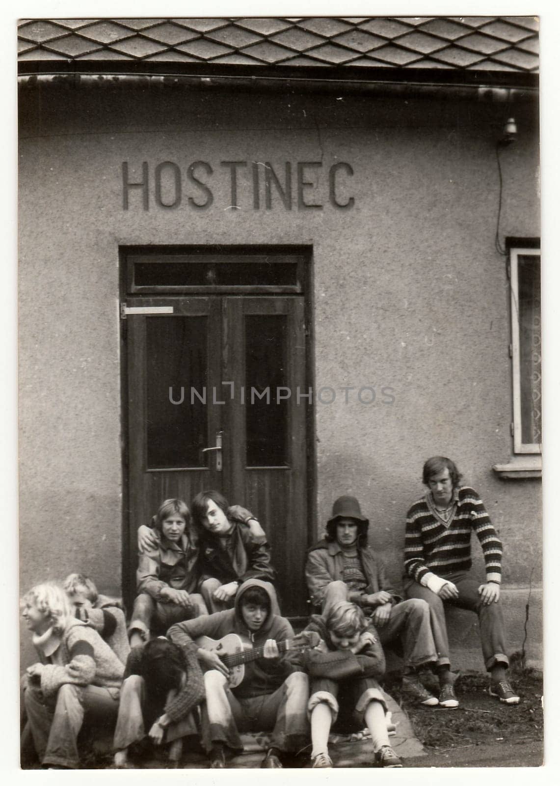 Vintage photo shows young hikers in front of the pub. One of them plays the guitar. Antique black white photo. by roman_nerud
