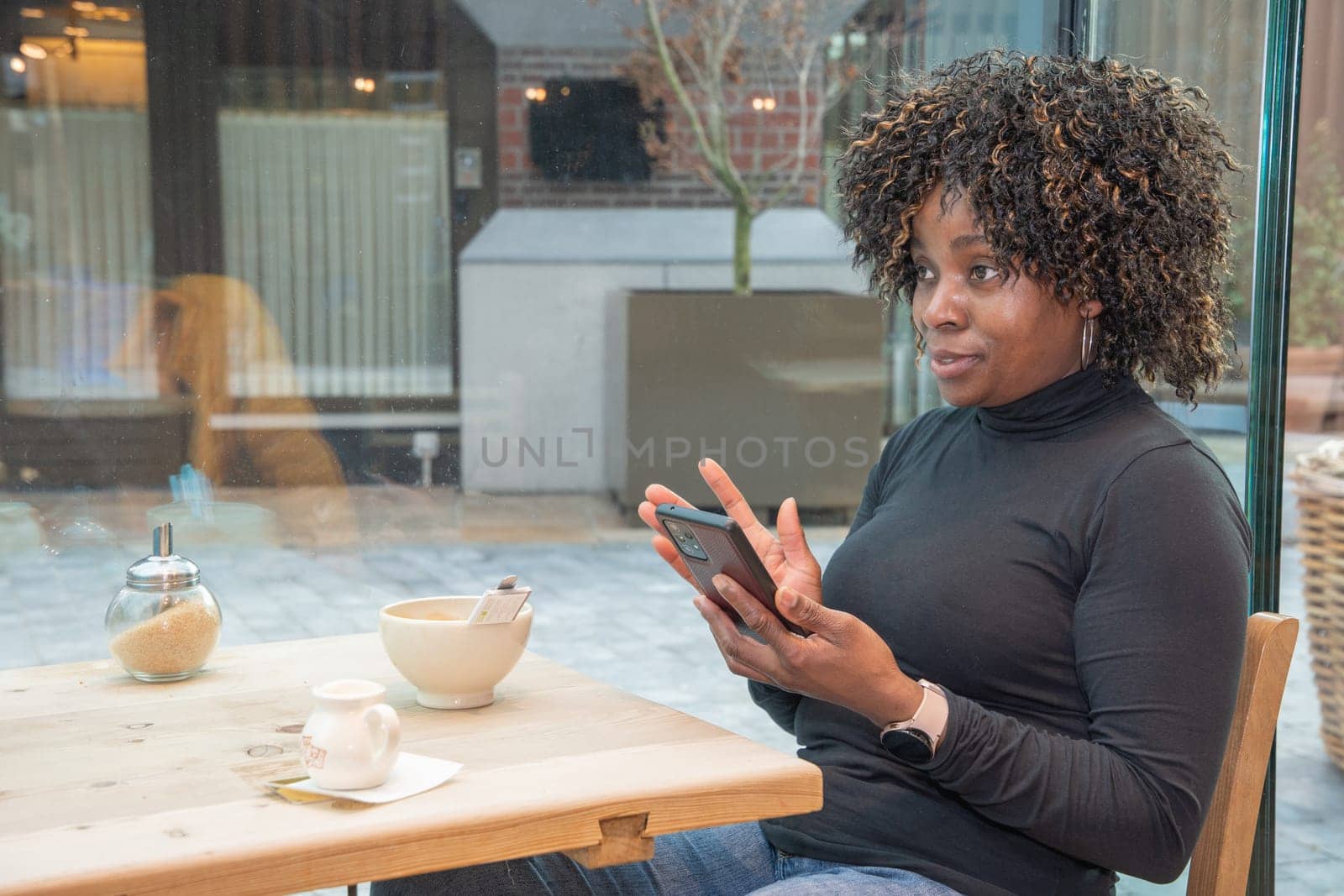 African american woman without makeup chats on smartphone in cafe,early morning by KaterinaDalemans