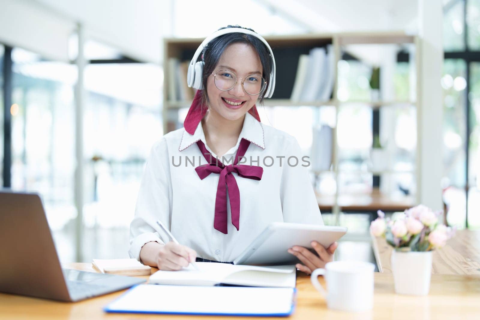 Portrait of a teenage Asian woman using a tablet, wearing headphones and using a notebook to study online via video conferencing on a wooden desk in library by Manastrong