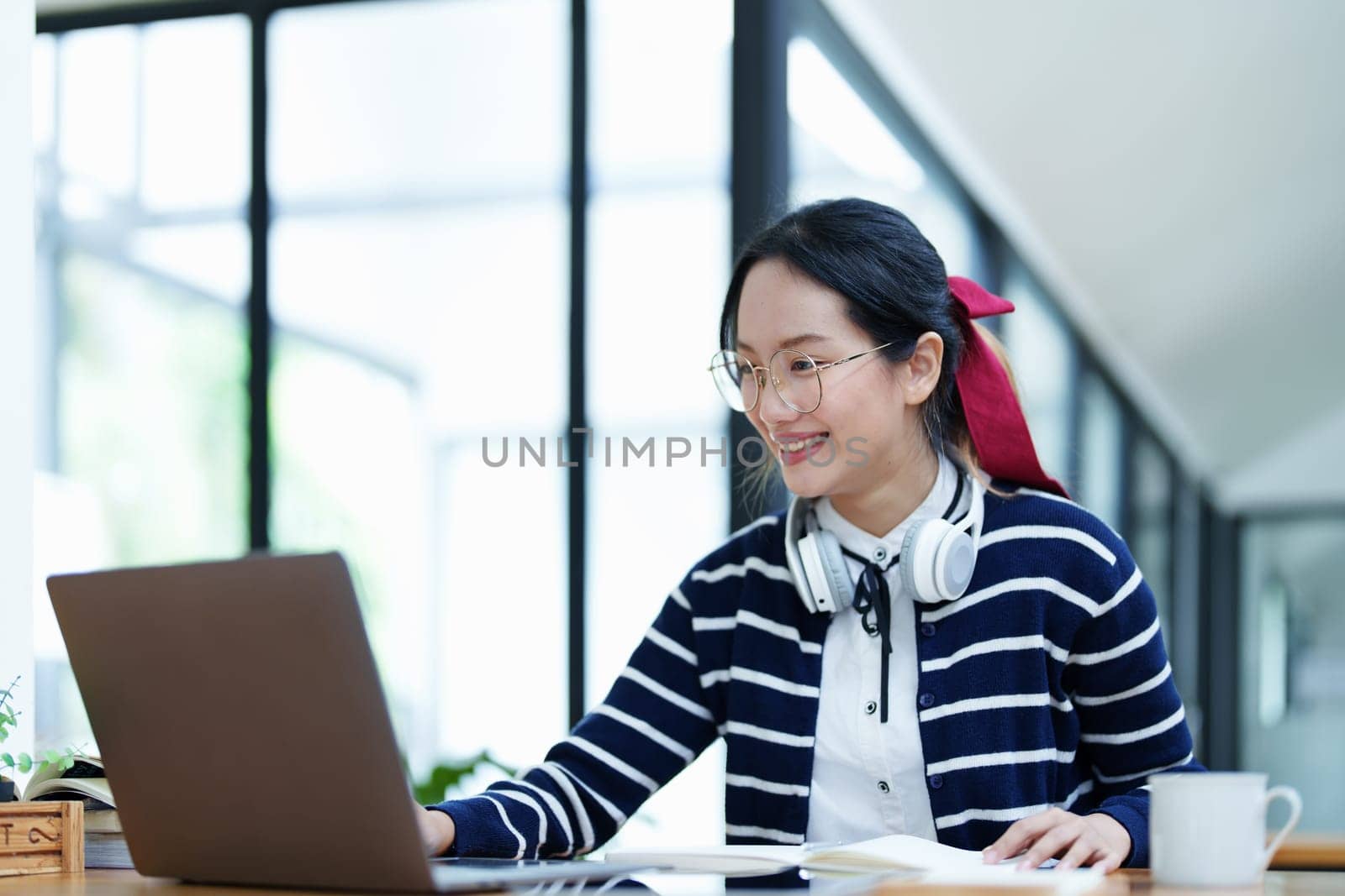 Portrait of a teenage Asian woman using a computer, wearing headphones and using a notebook to study online via video conferencing on a wooden desk in library.