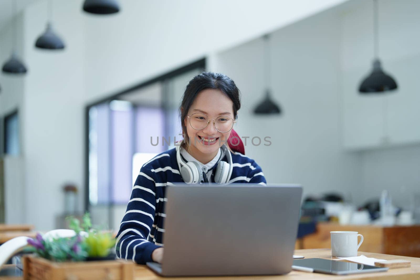 Portrait of a teenage Asian woman using a computer, wearing headphones and using a notebook to study online via video conferencing on a wooden desk in library.